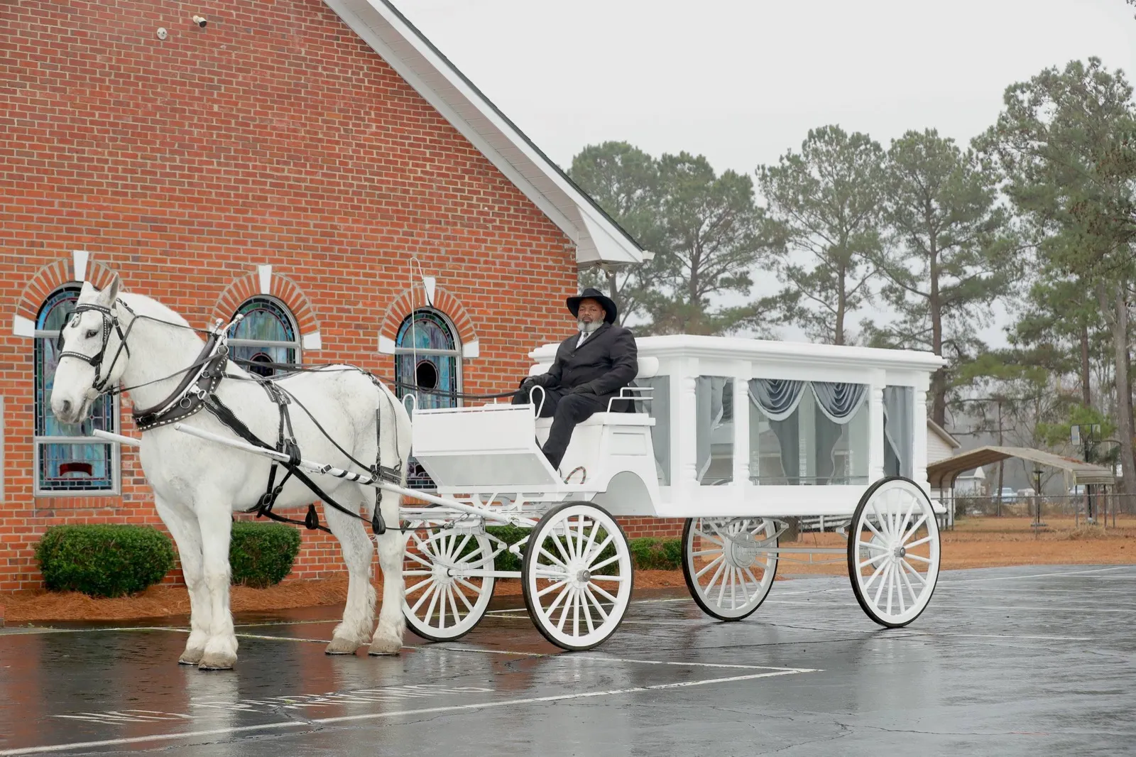White horse-drawn hearse carriage and white Percheron with a uniformed driver outside a brick church